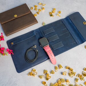 May include: A navy blue leather jewelry organizer with a pink watch band and a black bracelet. A brown leather wallet is also present. Gold-colored earring findings are scattered around the organizer.