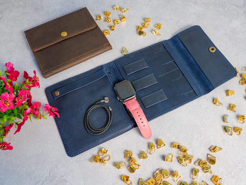 May include: A navy blue leather jewelry organizer with a pink watch band and a black bracelet. A brown leather wallet is also present. Gold-colored earring findings are scattered around the organizer.