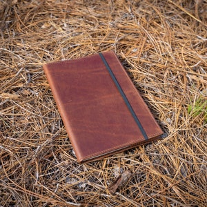 May include: A brown leather journal with a black elastic closure. The journal is lying on a bed of brown pine needles.