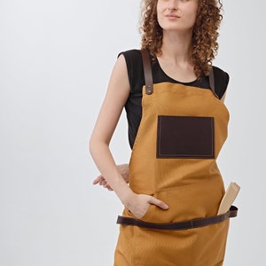 May include: A woman wearing a brown canvas apron with a brown leather pocket and a brown leather strap. She is standing with her hands in her pockets and looking up.