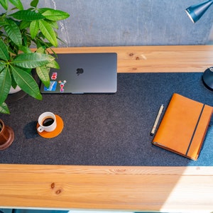 May include: A gray laptop computer, a brown leather notebook, a cup of coffee, and a desk lamp on a wooden desk with a gray felt desk mat.