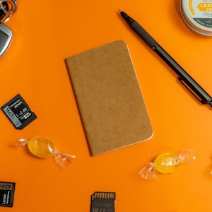 May include: A flat lay of various items on an orange surface, including a small brown notebook, a black pen, two SD cards, two yellow candies, and a small tin container with the text "Leather Cream".