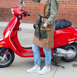 May include: A red Vespa scooter parked on a sidewalk with a person wearing a brown and green apron and blue jeans standing next to it.