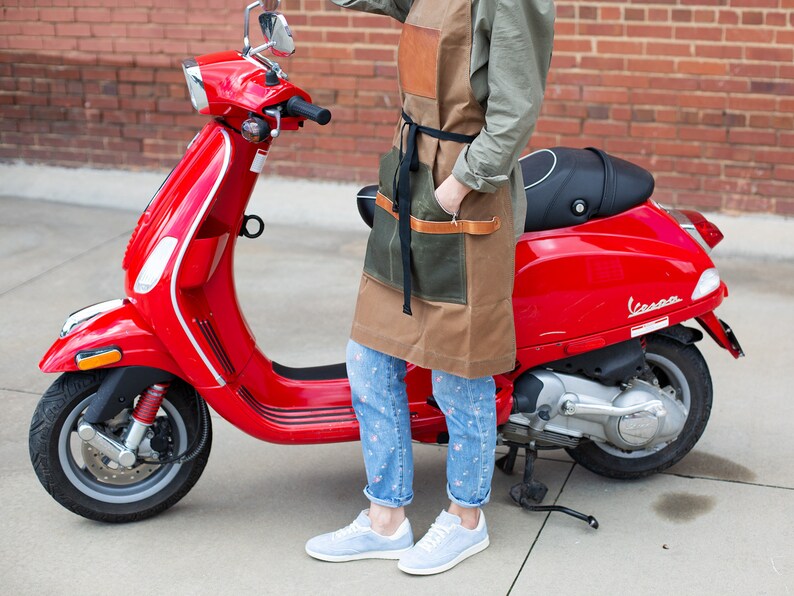 May include: A red Vespa scooter parked on a sidewalk with a person wearing a brown and green apron and blue jeans standing next to it.