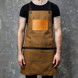 Puede incluir: Un delantal de lona marrón con correas de hombro de cuero marrón oscuro con remaches metálicos, un parche rectangular de cuero color canela claro en el pecho, una correa de cintura de cuero marrón oscuro y un bolsillo frontal grande con costuras.