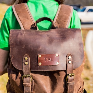 Puede incluir: Mochila de cuero y lona marr&oacute;n con herrajes de lat&oacute;n. La solapa presenta un parche de cuero con las iniciales "TAL". La mochila tiene correas ajustables y un asa superior. La mochila la lleva una persona con una camisa verde.