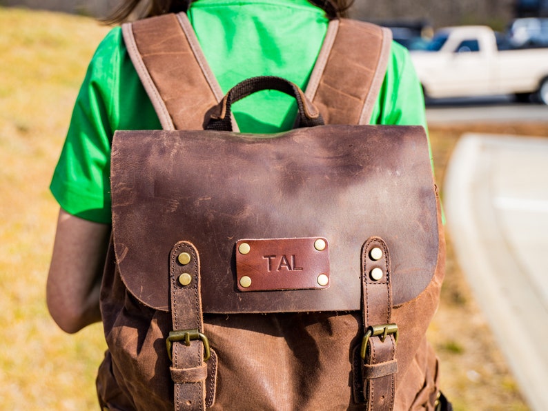 Puede incluir: Mochila de cuero y lona marr&oacute;n con herrajes de lat&oacute;n. La solapa presenta un parche de cuero con las iniciales "TAL". La mochila tiene correas ajustables y un asa superior. La mochila la lleva una persona con una camisa verde.