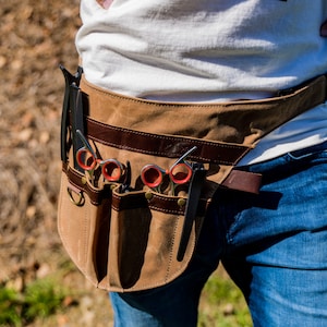 Puede incluir: Un cinturón de herramientas de lona marrón con múltiples bolsillos y un ribete de cuero. El cinturón se lleva alrededor de la cintura y contiene una variedad de herramientas de aseo, incluyendo tijeras y peines.