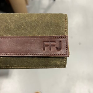 K&ouml;nnte beinhalten: Eine gr&uuml;ne Canvas-Geldb&ouml;rse mit einer braunen Lederklappe. Die Klappe hat die Initialen "FFJ" gepr&auml;gt.