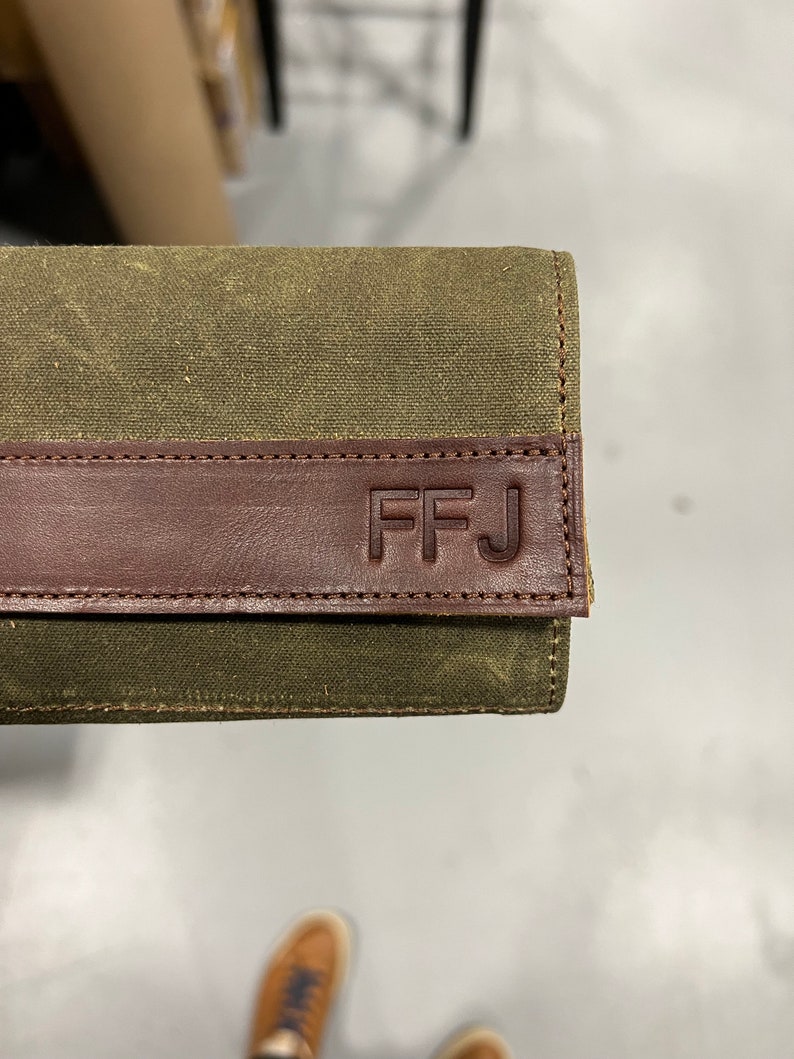 K&ouml;nnte beinhalten: Eine gr&uuml;ne Canvas-Geldb&ouml;rse mit einer braunen Lederklappe. Die Klappe hat die Initialen "FFJ" gepr&auml;gt.