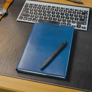 以下が含まれることがあります： 青い革張りのノートに黒いペンが乗っています。銀色のキーボードと茶色の革ストラップの腕時計も机の上にあります。ノートは閉じられています。