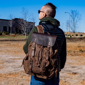 Puede incluir: Una mochila de lona encerada marr&oacute;n con detalles de cuero. La mochila tiene m&uacute;ltiples bolsillos, correas ajustables y un cierre de solapa. La mochila la lleva una persona con una sudadera con capucha verde y un chaleco negro, con gafas de sol.