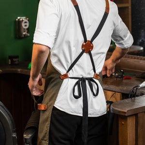 May include: A person wearing a white shirt and a green apron with brown leather straps. The apron has a black ribbon tied at the waist. The person is standing in a barber shop.