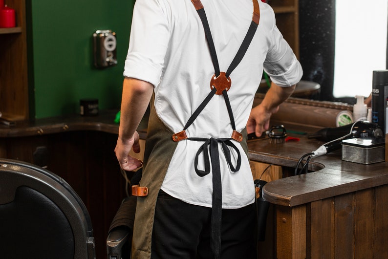 May include: A person wearing a white shirt and a green apron with brown leather straps. The apron has a black ribbon tied at the waist. The person is standing in a barber shop.
