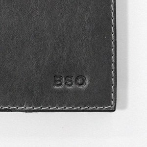 May include: Close-up of a dark gray leather item with the embossed letters "BSO". The item has a textured surface and white stitching along the edge. The corner of the item is visible against a white background.