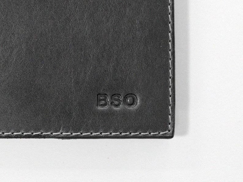 May include: Close-up of a dark gray leather item with the embossed letters "BSO". The item has a textured surface and white stitching along the edge. The corner of the item is visible against a white background.