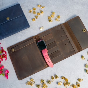 May include: A brown leather watch and jewelry organizer, open to display a pink watch. A navy blue leather pouch with a snap closure is next to the organizer. Gold-colored jewelry findings are scattered around the items.