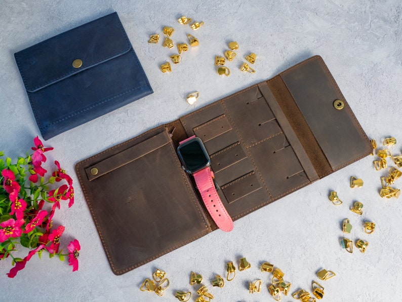 May include: A brown leather watch and jewelry organizer, open to display a pink watch. A navy blue leather pouch with a snap closure is next to the organizer. Gold-colored jewelry findings are scattered around the items.