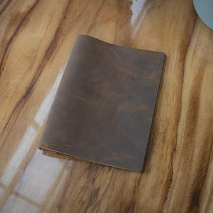 Op de afbeelding: Een bruin leren notitieboek ligt open op een houten oppervlak. Het notitieboek heeft een versleten, getextureerde uitstraling, met zichtbare nerf en subtiele kleurvariaties. De houtnerf van het oppervlak is prominent aanwezig.