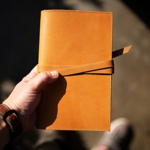 May include: A tan leather journal with a leather strap closure. The journal is closed and held in a person's hand.