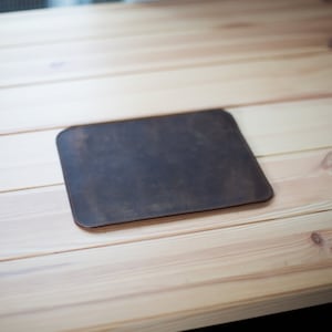 May include: A rectangular, dark brown leather desk mat with rounded corners. The mat is placed on a light-colored wooden surface. The leather has a distressed appearance with visible grain and a stitched border.