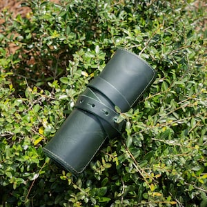 May include: A dark green leather watch roll with a strap closure. The cylindrical case is rolled up and secured with a matching strap. The watch roll is resting on a bed of green foliage.