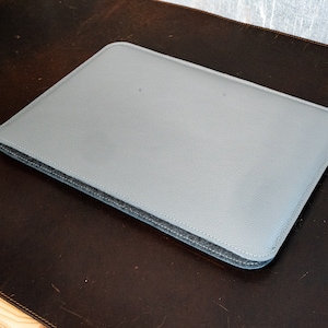 May include: A gray laptop sleeve with a stitched edge. The sleeve is made of a soft, textured material.