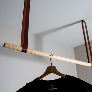 K&ouml;nnte beinhalten: Eine Holz-Kleiderstange, die an zwei braunen Lederriemen h&auml;ngt. Ein schwarzes T-Shirt h&auml;ngt an einem Holz-Kleiderb&uuml;gel an der Stange.