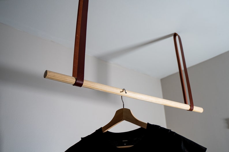 K&ouml;nnte beinhalten: Eine Holz-Kleiderstange, die an zwei braunen Lederriemen h&auml;ngt. Ein schwarzes T-Shirt h&auml;ngt an einem Holz-Kleiderb&uuml;gel an der Stange.
