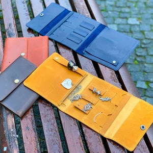 May include: An assortment of leather jewelry organizers in various colors, including brown, orange, and navy blue. One organizer is open, displaying necklaces, rings, and earrings. The organizers have snap closures and are designed for travel.