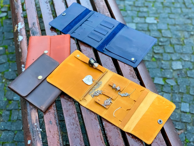 May include: An assortment of leather jewelry organizers in various colors, including brown, orange, and navy blue. One organizer is open, displaying necklaces, rings, and earrings. The organizers have snap closures and are designed for travel.