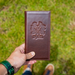 Puede incluir: Un diario encuadernado en cuero marrón con un logotipo en relieve dorado que representa un árbol y el texto "M.B.G. 1921".