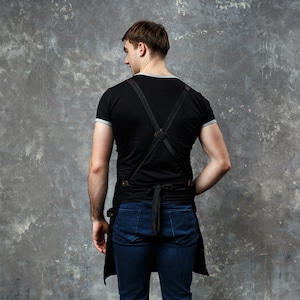 May include: A person wearing a black t-shirt with a black leather apron with straps. The apron has a long bib and a waist tie. The person is wearing blue jeans.