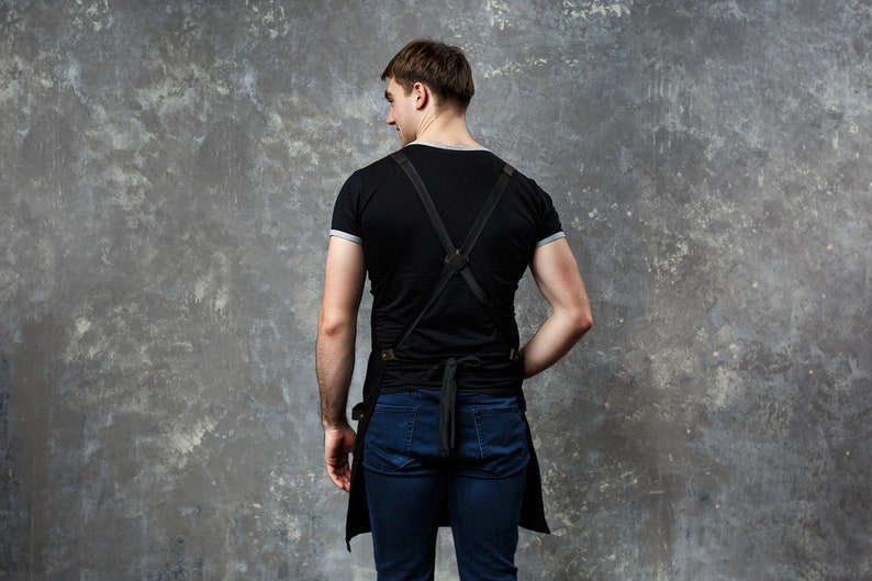 May include: A person wearing a black t-shirt with a black leather apron with straps. The apron has a long bib and a waist tie. The person is wearing blue jeans.