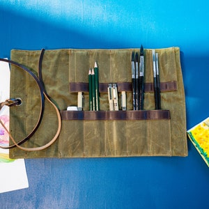 K&ouml;nnte beinhalten: Eine Canvas-Rolltasche mit einem braunen Lederriemen und mehreren Taschen zum Verstauen von K&uuml;nstlerbedarf. Die Tasche ist gef&uuml;llt mit Bleistiften, Stiften und Pinseln.