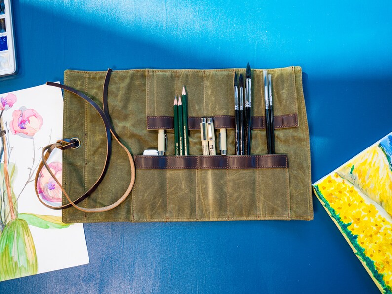 K&ouml;nnte beinhalten: Eine Canvas-Rolltasche mit einem braunen Lederriemen und mehreren Taschen zum Verstauen von K&uuml;nstlerbedarf. Die Tasche ist gef&uuml;llt mit Bleistiften, Stiften und Pinseln.