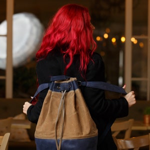 May include: A woman with long, bright red hair is wearing a black coat and a brown and blue canvas drawstring backpack. The backpack has a long, adjustable strap.