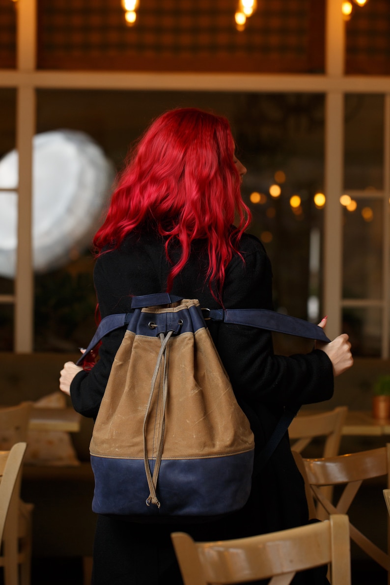 May include: A woman with long, bright red hair is wearing a black coat and a brown and blue canvas drawstring backpack. The backpack has a long, adjustable strap.
