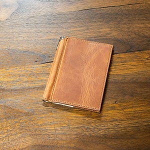 Puede incluir: Una cartera de cuero marrón con un clip de dinero de metal. La cartera está cerrada y se encuentra sobre una superficie de madera.