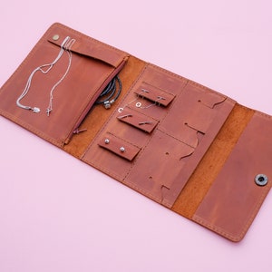 May include: Open, brown leather jewelry organizer with multiple compartments for necklaces, earrings, and bracelets. The organizer is open, revealing various pieces of jewelry. The background is a solid pink color.