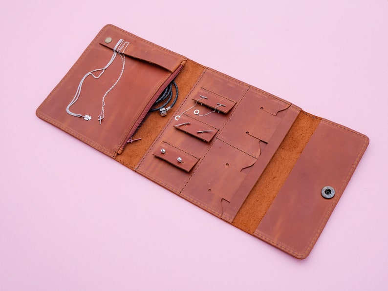 May include: Open, brown leather jewelry organizer with multiple compartments for necklaces, earrings, and bracelets. The organizer is open, revealing various pieces of jewelry. The background is a solid pink color.
