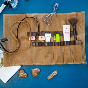 May include: A brown canvas roll-up case with multiple pockets for storing makeup and beauty products. The case is open and shows a variety of items, including a brush, a tube of face cream, and a small bottle of lotion. The case is on a blue surface.