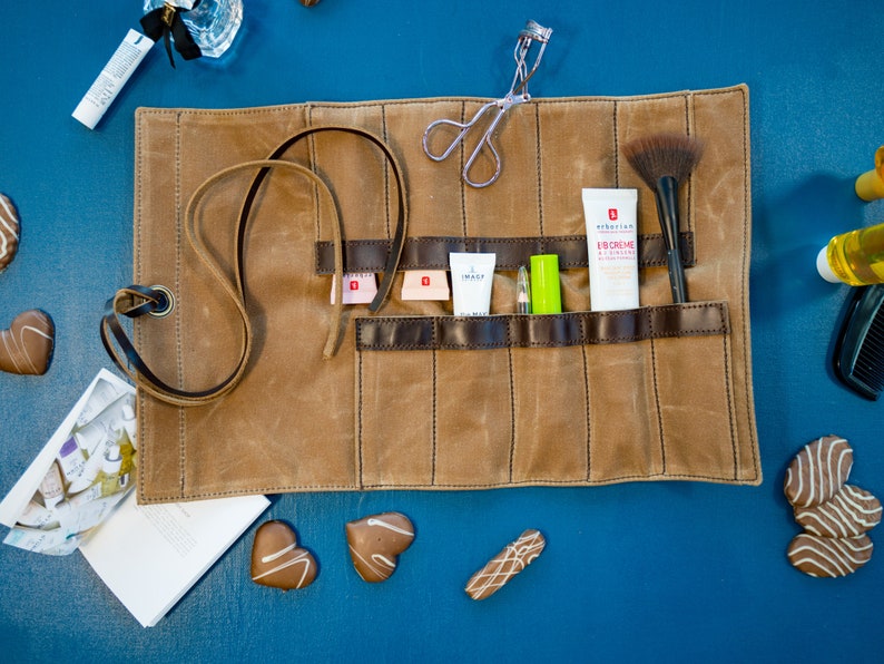 May include: A brown canvas roll-up case with multiple pockets for storing makeup and beauty products. The case is open and shows a variety of items, including a brush, a tube of face cream, and a small bottle of lotion. The case is on a blue surface.