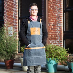 May include: A man wearing a gray denim apron with a brown leather patch that says "Radish" in cursive. The apron has a brown leather strap and two pockets. The man is standing in front of a brick building with windows.
