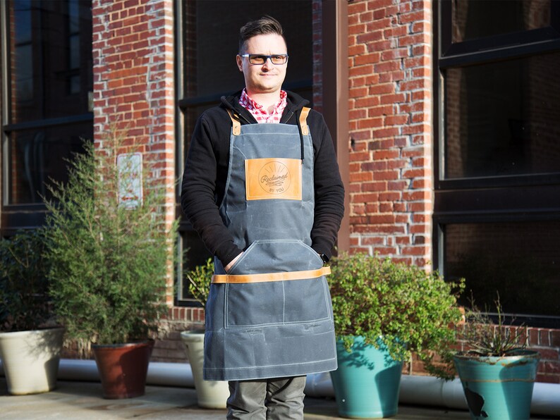 May include: A man wearing a gray denim apron with a brown leather patch that says "Radish" in cursive. The apron has a brown leather strap and two pockets. The man is standing in front of a brick building with windows.