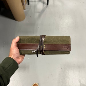 K&ouml;nnte beinhalten: Eine gr&uuml;ne Canvas-Rolltasche mit einem braunen Lederriemen und einer Messing-Schnalle. Die Tasche hat die Initialen "FFJ" auf dem Lederriemen gepr&auml;gt.