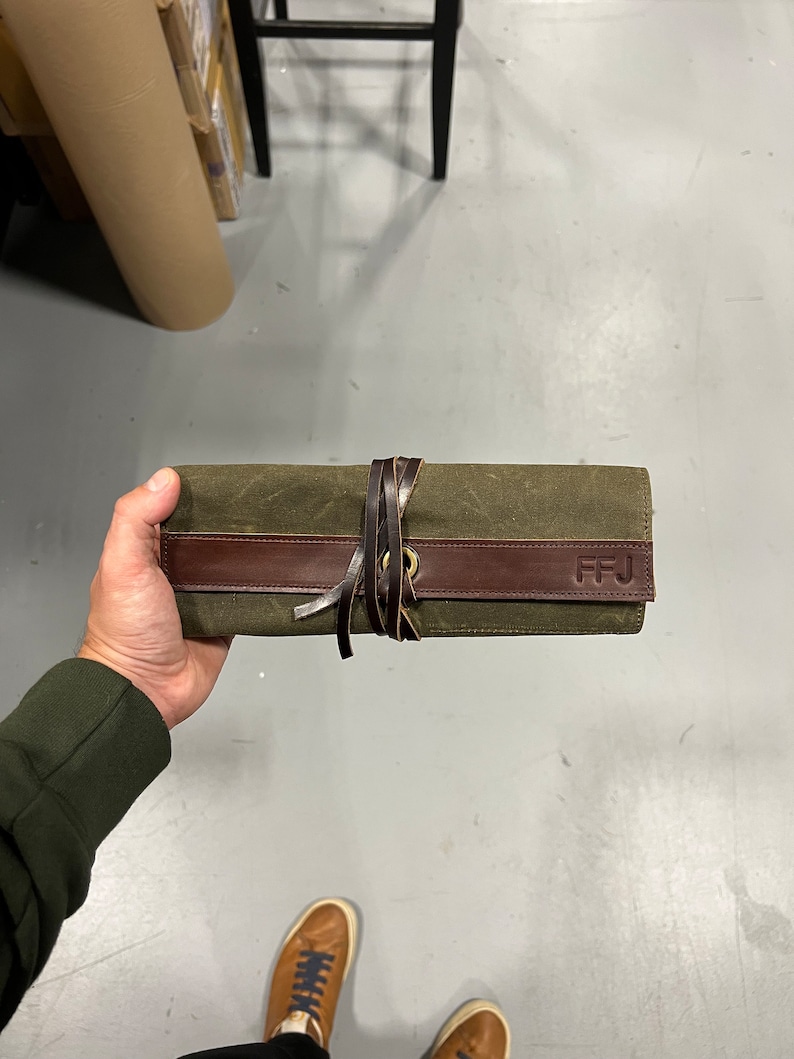 K&ouml;nnte beinhalten: Eine gr&uuml;ne Canvas-Rolltasche mit einem braunen Lederriemen und einer Messing-Schnalle. Die Tasche hat die Initialen "FFJ" auf dem Lederriemen gepr&auml;gt.