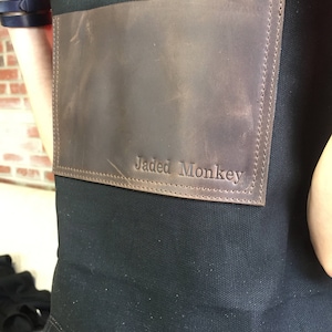 May include: A black canvas apron with a brown leather patch that says "Jaded Monkey" in white stitching.