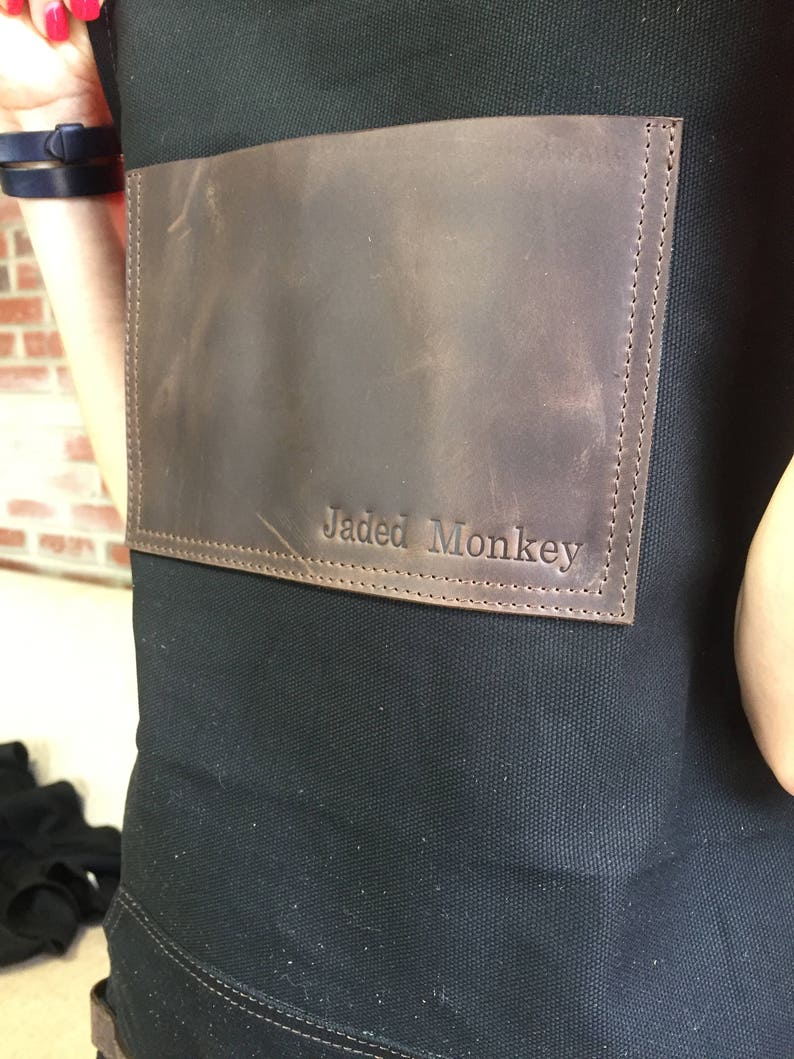 May include: A black canvas apron with a brown leather patch that says "Jaded Monkey" in white stitching.