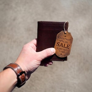 May include: A dark brown leather wallet with a matching leather tag. The tag reads "CHESTNUT SPECIAL SALE LIMITED TIME" and is attached with a string. The wallet is held in a hand, with a brown leather watch visible.